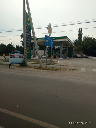 The image captures a gas station in Sokuluk, Kyrgyzstan, with a prominent price board displaying fuel costs. A mock police car, likely a promotional display, is parked to the left of the frame. The gas station, branded "Bishkek Petroleum," features multiple pumps under a covered canopy. The surrounding environment appears to be a moderately vegetated area with trees lining the background. The sky is overcast, suggesting possibly cloudy weather. A street and pavement are visible in the foreground and to the right. The timestamp "19.06.2026 11:51" is embedded in the bottom right corner of the image.