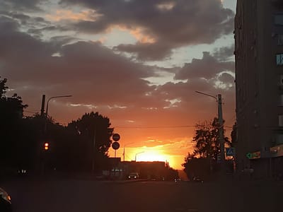 The image depicts a sunset in the city of Shakhty, Russia. The sky is filled with dramatic clouds, transitioning from deep grays and purples to vibrant oranges and yellows near the horizon where the sun is setting. Silhouetted against the bright sky are trees, streetlights, traffic signals, and the side of a tall apartment building. The street level shows a road with a few cars and signs, including a pharmacy with a green cross emblem. The overall impression is a dusky urban scene illuminated by the warm glow of the setting sun.