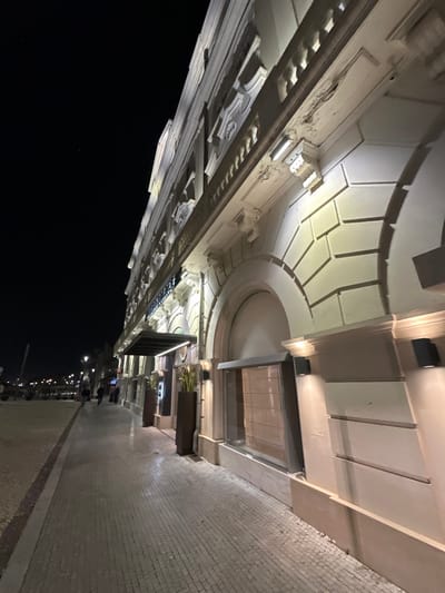 The image depicts the illuminated facade of the Hotel Aveiro Palace at night, located on Rua Dr. Vítor Círio in Aveiro, Portugal, facing the Central Canal. The multi-story building features a light-colored, possibly cream, neoclassical exterior with ornate architectural elements including decorative balustrades, cornices, pilasters, and multiple arched recesses on the ground floor. The facade is extensively lit by warm white upward and downward-facing wall-mounted fixtures and linear strip lighting, highlighting its details. Minor discoloration and surface wear are visible on some upper architectural elements.
At street level, a central entrance is situated beneath a dark, cantilevered canopy. This entrance is flanked by two tall, dark rectangular planters containing green foliage. To the right of the entrance, an adjacent arched recess contains a large window with an internal rolled blind. The immediate foreground comprises a patterned cobblestone sidewalk, which extends into the mid-ground. Two indistinct human figures are visible walking away from the camera on this sidewalk. To the left of the sidewalk, a darker area consistent with a street or promenade is visible, featuring distant streetlights and blurred lights suggesting other structures or slow-moving vehicles. The sky is dark black, indicating nighttime. Silhouetted vegetation, consistent with trees, is present in the distant background on the left.