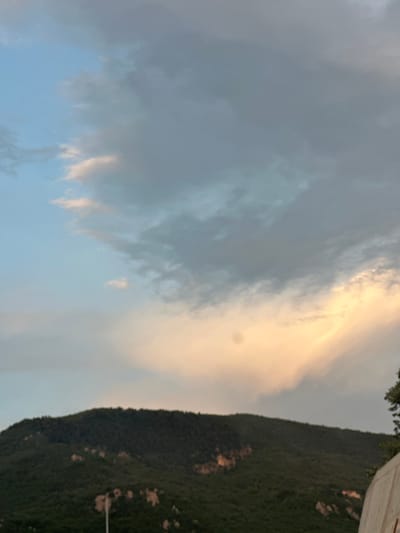 This media file captures a scenic view of a mountain range and the sky. The mountain is covered in dark green vegetation, with some lighter patches suggesting rocky outcrops or sparse vegetation. The sky is a blend of soft blues and grays, with dramatic clouds that are illuminated with warm, golden light, indicating either sunrise or sunset. The lighting suggests a serene and possibly tranquil atmosphere. The image appears to be taken outdoors, given the presence of the mountain and sky. There are no visible people or specific objects that stand out prominently, making the natural landscape the primary subject. The location context suggests this scene is in Lastva Grbaljska, Montenegro. The time of day is likely during twilight due to the sky's colors.