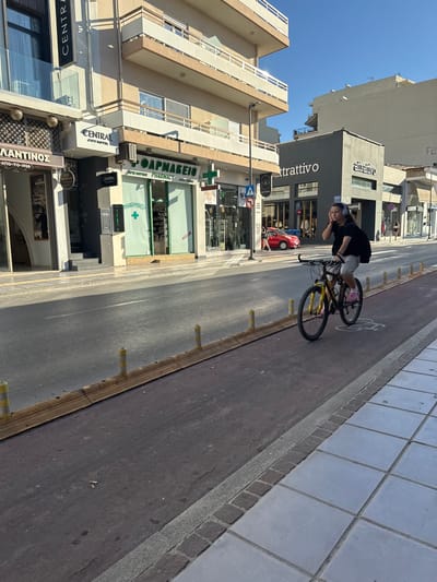 The image captures a street scene in Chania, Greece, likely on a sunny day. In the foreground, a person is riding a bicycle on a dedicated bike lane that runs alongside the road. The cyclist, wearing a black t-shirt and light-colored shorts, has headphones on and appears to be touching their ear. The bicycle is dark-colored with yellow accents and pink shoes are visible on the rider's feet.
The street itself is paved, with a red-colored bike lane separated from the main road by yellow bollards and a wooden barrier. On the right side of the road, a sidewalk with large square tiles is visible.
The background features buildings lining the street. On the left, a building with the sign "CENTRAL CITY HOTEL" is prominent, with shops below. To the right, other commercial establishments are visible, including one with a green facade and the word "PHARMACY" in English and Greek. Further down the street, more buildings and shops are present, with one displaying the sign "attrattivo". A red car is parked on the street. The sky is clear and blue. The overall scene depicts a typical urban environment in Chania.