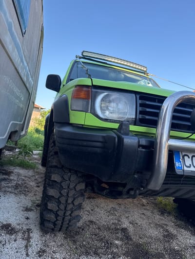 A low-angle, close-up shot captures the front left side of a bright green Mitsubishi Pajero off-road vehicle parked outdoors. The vehicle features a rugged, textured paint finish and is equipped with oversized, knobby tires, a prominent chrome bull bar, and a long LED light bar mounted above the windshield. A Bulgarian license plate, partially visible, reads "BG CO". The vehicle is positioned next to a large, light-colored trailer or container, its side also visible in the frame. The environment appears to be a somewhat unkempt outdoor area, with gravel and patchy vegetation on the ground. In the background, a residential building and trees are discernible under a clear blue sky. The scene is consistent with a location in Lesnovo, Bulgaria, a rural area known for its proximity to nature and outdoor activities, suggesting the vehicle is prepared for or has been used for off-road excursions. No people are present in the image.