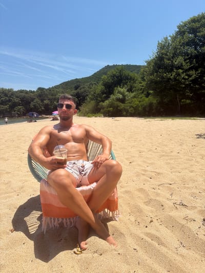 The image displays a man seated on a sandy beach in Ολυμπιάδα, Greece, under a clear blue sky with visible contrails. The man has short dark hair, a beard, and wears dark aviator sunglasses. He is shirtless, revealing a muscular, tanned physique with a small piercing on his left nipple and a faint mark on his upper left chest. He wears light-colored swim shorts with a floral pattern. He is seated in a chair constructed with light blue and white cords, over which an orange and white striped towel is draped. His legs are crossed at the ankles. In his right hand, he holds a clear plastic cup with a lid and straw, containing a dark liquid. The background features a body of water leading to a shoreline with additional individuals and colorful beach umbrellas (blue and pink) in the distance. Beyond the water, lush green hills covered in dense trees rise under the bright sunlight, which casts distinct shadows of the man and chair onto the golden sand.