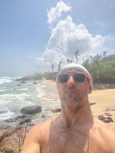 This is a selfie taken by a man on a beach in Midigama, Sri Lanka. The man is wearing sunglasses and a white head covering. The beach has sand, rocks, and palm trees. The ocean is visible with waves crashing on the shore. The sky is blue with white clouds. The time of day appears to be daytime, and the weather seems sunny. The image is oriented vertically.