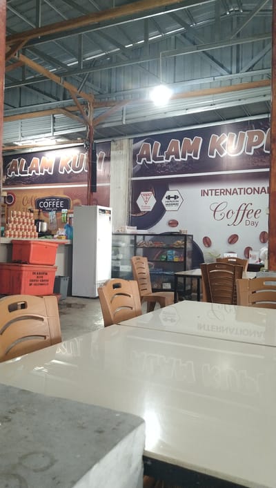 The image depicts the interior of a coffee shop. In the background, banners with "ALAM KUP" and "INTERNATIONAL COFFEE DAY" are visible. There is a refrigerator, a glass display case filled with items, and several tables and chairs. Some of the chairs are made of light brown plastic, while others appear to be wooden. In the foreground, a white reflective table is partially visible, with a gray object to its left. Stacks of orange plastic containers are also present in the left side of the image. The overall environment suggests a casual, possibly outdoor or semi-outdoor dining or resting area within a commercial establishment. The presence of a refrigerator and display case indicates food and beverage service. The lighting suggests it is daytime, with overhead artificial lights illuminating the space. The signs and context point towards a coffee-related business.