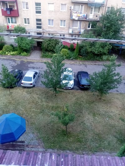 The image depicts an outdoor scene adjacent to a multi-story apartment building, likely in Tiachiv, Ukraine. Several cars are parked in a lot. A blue umbrella is open and resting on a brick pathway. The ground is covered in dry grass, with some scattered trees and bushes. The apartment building has multiple windows and balconies, some with laundry hanging out. The overall impression is a residential area in a town or city.
