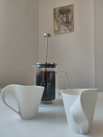 The image depicts a still life composition on a white surface, likely a table. In the foreground, two white ceramic cups with a unique, sculptural design are placed. The cups have a smooth, matte finish and their handles are integrated into their flowing forms. Behind the cups, a glass French press filled with dark liquid, presumably coffee, is centrally positioned. The French press has a silver lid and plunger mechanism. The background consists of a lightly textured, off-white wall. Hanging on the wall, slightly off-center to the right, is a small framed painting with a golden frame. The painting appears to be a landscape with an olive tree, stone steps, and a building. The overall lighting is soft and diffused, suggesting an indoor setting. There is no indication of the specific location within Zrenjanin, Serbia, as the image lacks any identifiable landmarks or contextual clues specific to that city.