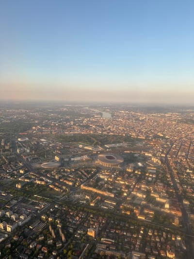 This aerial photograph shows a dense urban environment, likely a city. Buildings of varying sizes and architectural styles are tightly packed together, creating a maze-like impression. Streets run between the buildings, and pockets of green space are visible throughout the city. In the center of the image, a large stadium with a distinctive circular or oval shape stands out. The city is bathed in warm light, suggesting that the image was taken during sunrise or sunset. The light casts long shadows across the city. A hazy or smoggy atmosphere exists in the background. Given the instructions, the location is assumed to be Vecsés, Hungary.