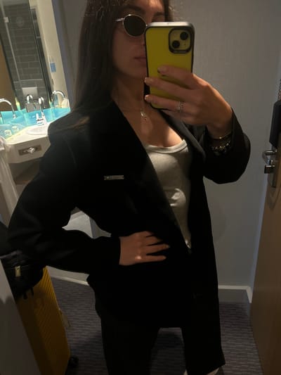 An individual is captured in a mirror selfie, holding a yellow-cased smartphone to take the picture. The person, likely a woman, has long dark hair and is wearing round, dark-lensed sunglasses. She is dressed in a black blazer or jacket over a light grey ribbed tank top. Accessories include a ring on her right ring finger, bracelets on her left wrist, and a delicate silver necklace.

The setting appears to be an indoor room, possibly a hotel room given the visible elements. Reflected in the mirror behind the individual is a bathroom area with two modern faucets and what looks like a tiled wall. The room itself features a textured grey carpet or flooring, and a partially visible yellow pleated suitcase can be seen towards the bottom right of the main image. The lighting in the room is dim, creating some shadows and highlights on the clothing.

Notable details include the yellow phone case, which has "WORDELIER" or a similar stylized brand name visible on its side. A small, rectangular silver tag with indistinct, stylized text (possibly a brand name starting with 'V' or 'M') is attached to the black blazer near the lapel. The overall scene suggests a casual, possibly travel-related, moment. The location context provided is Dortmund, Germany.