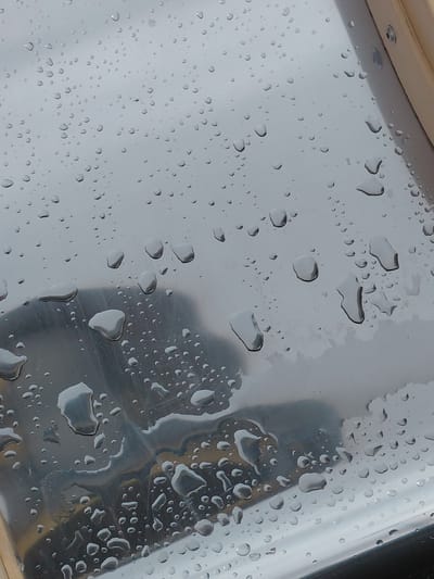 A close-up, high-angle shot captures numerous water droplets of varying sizes on a translucent, reflective surface, likely a window or pane. The droplets range from tiny, distinct spheres to larger, irregularly shaped pools, suggesting recent or ongoing rain. Through the water and the surface, a soft, light grey, blurred background is visible, indicative of an overcast sky or distant, indistinct surroundings.
In the upper left corner, a sliver of a light-colored, possibly wooden, frame or trim is present, secured by a metallic screw or rivet. This detail hints at an architectural feature such as a window, skylight, or a vehicle's viewport. The right and lower portions of the image feature darker, abstract shapes and reflections, possibly originating from objects or structures on the side of the surface not facing the light background. The lighting is diffuse, consistent with an overcast day. No people, specific activities, or discernible text are visible. The image conveys the natural phenomenon of rain observed through a built structure, set against the backdrop of Nadym, Russia.