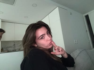A woman with brown hair and red nail polish poses with pursed lips and a finger to her mouth. She wears a black top and looks into the camera. Behind her, there are white cabinets and countertops, suggesting a kitchen area. In the background, a man can be seen faintly near the kitchen. To her right, a door and a grey blanket are visible. The room is brightly lit with recessed lighting in the ceiling.
