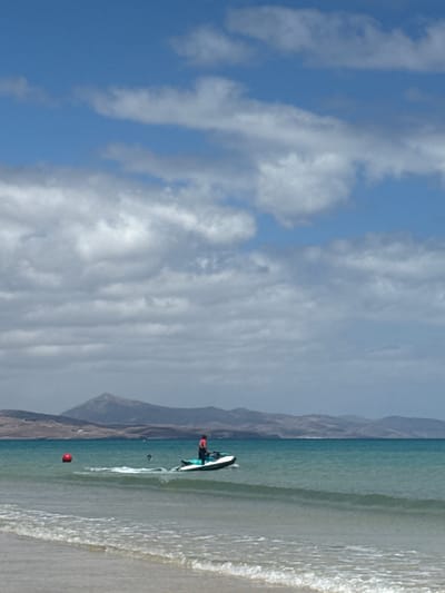 The image shows a beach scene in Pájara, Spain. A person is on a stand-up paddleboard in the turquoise water, near the shoreline. In the water, there is a red buoy floating. Mountains are visible in the background. The sky is blue with many white clouds. The beach has tan sand and small waves are crashing on the shore.