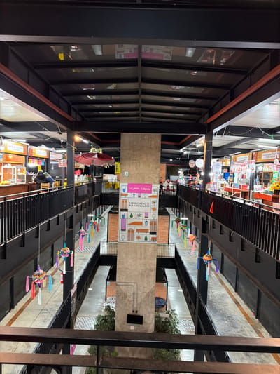 The image is of a covered market. There are colorful stalls and signs with food and other products. The market is decorated with bright paper flowers and ribbons, creating a festive atmosphere. Metal beams and a grid-like structure support the roof, casting shadows on the stalls below. There are windows on the left. A large sign labeled "MERCADO DE PATZCUARO" hangs in the middle of the market.