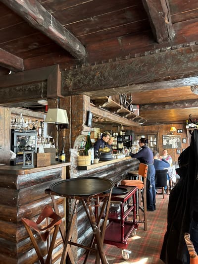 The image displays the rustic interior of a bar or restaurant, characterized by prominent dark, heavy wooden beams and paneling throughout, creating a traditional, cabin-like aesthetic. The floor is covered with a plaid-patterned carpet.
Several individuals are visible, suggesting the establishment is operational. Two men, likely bartenders, are positioned behind a long, sturdy wooden bar counter. One, with blonde hair, stands upright, while another, with dark hair, leans forward, appearing to be working or preparing drinks. In the background, two customers are seated at a table: one man wears a plaid shirt, and another individual is partially obscured.
The bar counter is well-stocked with bottles, including a large champagne bottle in an ice bucket, various glasses, and bar equipment such as a coffee machine and a golden cup or trophy. Above the counter, shelves hold additional bottles. Walls are decorated with framed black and white photographs. A small square lamp is attached to a wooden beam, providing warm, ambient lighting.
In the foreground, a large, dark, circular tray rests on a wooden stand. To the right, a wooden trolley with red upholstery is visible. The heavy wooden construction, including exposed beams and joinery, contributes to the establishment's robust and antique feel. A small, black rectangular device, possibly a menu or point-of-sale terminal, is on the bar counter, but its text is illegible. The scene captures typical bar activities within a cozy and inviting atmosphere.