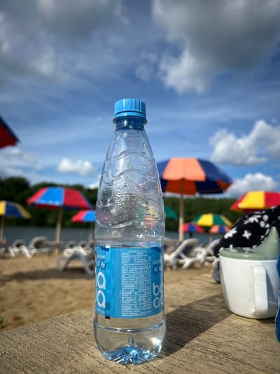 In the foreground, a clear plastic bottle of water with a blue cap stands on a weathered wooden surface. The bottle has a blue label with white text and the logo "BonAqua". Water droplets are visible on the exterior of the bottle. To the right of the bottle, a white sippy cup with a green lid is partially visible, also with water droplets. In the background, a sandy beach is lined with lounge chairs and colorful beach umbrellas in red, blue, orange, and yellow. The water appears calm, and trees and a hill are visible on the opposite bank of the body of water. The sky is partly cloudy with blue sky visible between the clouds. The scene depicts a relaxing day at a beach or lakeside, possibly in the city of Yubileynyy, Russia, given the labeling on the bottle. There are no people clearly visible in the image.