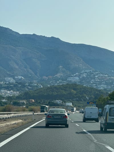 This image captures a scenic view from a highway in Altea, Spain, likely during the daytime with clear weather. The perspective is from inside a vehicle, looking forward and slightly to the left, suggesting traffic is moving along the road.
The primary subjects are vehicles on a highway, including a dark-colored car in the foreground with license plate "2774 GDJ", followed by white vans and trucks. One van has the license plate "1758 NCK". Overhead signs are visible, with text that includes "AP-7", "639", and "A 621", indicating this is a major road.
The setting is a dramatic mountainous landscape bordering the highway. The mountains are rugged and covered with dense green vegetation and scattered white buildings, characteristic of a Mediterranean coastal town. The buildings cling to the steep hillsides, creating a picturesque and densely populated urban area nestled within nature. The sky is a light, clear blue, suggesting good visibility and a pleasant day. The time of day appears to be midday or early afternoon, given the bright light and clear shadows cast on the road.
The overall mood is one of travel and observation, with the traffic on the highway providing a sense of movement against the static beauty of the mountains and the town. The combination of the road, vehicles, and the stunning natural and urban landscape creates a typical scene of travel through a developed coastal region.