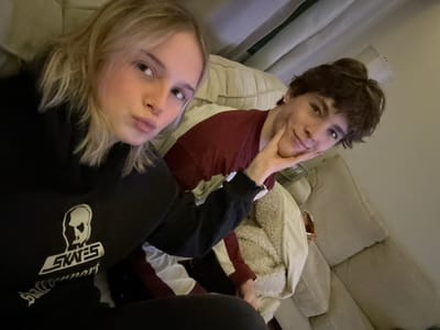 Two individuals are posing for a selfie while lying on a couch. One, with dark hair and a red sweater, has a hand gently touching their face. The other, with blonde hair and wearing a black hoodie with a distinctive white logo, is positioned closer to the camera. They are both looking towards the camera. The couch is beige, and a white jacket is visible in the background. The photo is taken at an unusual angle.