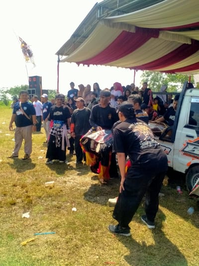 The image depicts an outdoor cultural gathering in Celep, Indonesia, under clear skies. A large, temporary canopy with alternating red and cream-colored stripes covers the right side of the frame. Beneath the canopy and extending into the open, a crowd of approximately 30-40 individuals, primarily adults, is visible; many are observing the central activity.
In the mid-ground, a central group of individuals is focused on what appears to be a performance or ceremonial preparation. A person wearing a black t-shirt with a gold-colored graphic on the back, reading "seni jaran..." (likely "Seni Jaranan") and "Sindurging...," is bent forward, interacting with an elaborate, colorful object. This object resembles a stylized horse head or mythical creature, featuring intricate patterns, prominent ears, and red and yellow tassels or mane. To the left, another man in a black t-shirt and a black-and-white checkered sarong-like garment observes, while a third man in a dark t-shirt and light pants stands further left.
To the far right, the rear section of a white pickup truck, marked with orange and black graphics, serves as a makeshift platform. At least two traditional Indonesian drums (kendang or similar Gamelan instruments) are visible in the truck bed, with one individual seated nearby, suggesting a musical performance component.
In the background to the left, a tall pole bears a white flag adorned with a dark, stylized emblem, likely an eagle or Garuda-like figure. A large black speaker stack is positioned near the flag. The ground consists of dry, sparse grass with scattered pieces of white debris and a blue plastic item visible in the foreground. Strong shadows cast by the people and objects indicate bright daylight. The distant background on the left shows an open, undeveloped field under a bright sky.