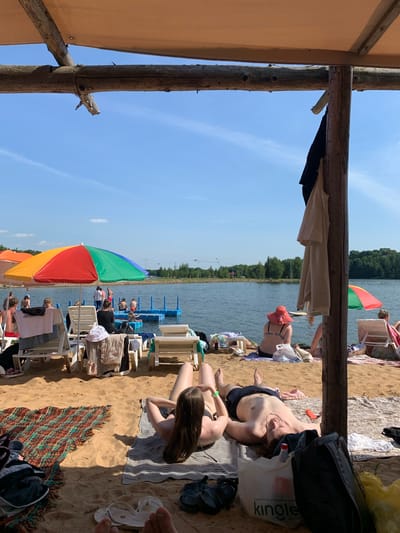 The image depicts a sandy beach area adjacent to a body of water, likely a lake or reservoir, on a sunny day. In the foreground, two individuals are lying on towels on the sand, sunbathing. One person, with long brown hair, is lying on their side, while the other, a shirtless male, is lying on their back. Beach chairs, umbrellas, and bags are scattered around the sand. Further back, several other people are visible on the beach and in the water, some near a blue floating platform. The water is calm, and the background shows a treeline. The overall scene suggests a recreational area, possibly within the Шчомысліцкі сельскі Савет region of Belarus, given the context. A wooden support structure for a canopy is prominent in the right foreground, with clothing hanging from it.