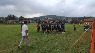 The media file depicts a football (soccer) match or training session taking place on a grassy field in a rural Indonesian setting, likely Panjalu, under an overcast sky.
In the center of the frame, a group of approximately fifteen young men or teenagers, dressed in black football jerseys, are gathered in a tight huddle. Their jerseys display numbers such as "28," "16," "77," and "2," along with text like "FC" and "MREZ." One player in an orange jersey stands slightly apart from this central group. To the far right of the frame, a different group of individuals, some wearing yellow jerseys, is visible further down the field, suggesting an opposing team or a group of substitutes. A person in a white collared shirt and light-colored pants is seen walking away from the camera in the foreground, towards the huddle.
The scene is set against a backdrop of traditional village houses with varied architecture and colors, nestled amongst lush tropical vegetation including palm trees. Rolling green hills or mountains dominate the distant background. The sky is completely overcast with thick grey clouds, indicating a cloudy day, possibly late afternoon given the diffused light and absence of harsh shadows. Orange and red ribbons or tape partially obscure the view on the right edge of the foreground, suggesting boundary markers or decorations for the event. A few spectators, including children, are visible on the sidelines, with some seated on chairs. The overall atmosphere suggests a community sporting event or practice session.