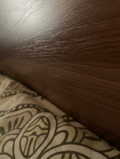 The image shows a close-up view of a brown wooden headboard of a bed, with a patterned bedsheet in the foreground. The wood grain is visible, and there are highlights reflecting off the polished surface. The bedsheet has a repeating abstract design in beige and dark grey. The lighting suggests an indoor setting. The image does not provide any information about the city of Oktyabrskoye, Russia, or any specific context beyond the immediate objects depicted.
