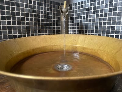 The image displays a close-up, eye-level view of a circular, golden-toned metal vessel sink with water actively flowing into it from a vertical, minimalist faucet. The sink exhibits an aged or patinated finish, with visible water droplets on its interior rim and standing water at its base, surrounding a circular metal drain. Directly behind the sink, a wall is tiled with small square mosaic tiles in varying shades of dark gray and black, arranged in a grid pattern with light-colored grout. There are no individuals present in the scene. The image provides no visual information to confirm the specific location within Juta, Georgia.