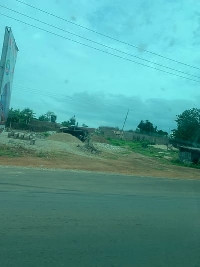 The image shows a street scene in Laminga, Nigeria. The road is paved and runs vertically along the right side of the frame. To the left of the road is a dirt shoulder and then a construction site. The construction site has piles of dirt and gravel, stacks of building blocks, and several unfinished structures with exposed rebar. There are trees and vegetation in the background, some of which appear to be banana trees. A utility pole with wires is also visible. In the far left of the image, a portion of a large banner is visible, with some text and logos. The sky is overcast and appears to be a pale blue or gray, suggesting it might be daytime with cloudy weather. There are no people clearly visible in the image, though there appears to be a figure in the background near the construction shed. The scene depicts ongoing construction and a somewhat rural or developing environment.