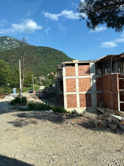 The image depicts a construction site in Kavač, Montenegro, with a partially built multi-story brick and concrete structure dominating the foreground and midground. The building appears to be in an early stage of construction, with exposed concrete beams, wooden scaffolding, and brick walls in place. Debris, including rocks and wood, is scattered around the base of the building and on the unpaved road leading to it. A portable toilet is visible to the left of the road. In the background, a lush green hillside with scattered houses and trees rises, under a bright blue sky with scattered white clouds. A single, bare tree stands near the portable toilet. The overall scene suggests ongoing construction activity in a semi-rural, hilly environment.