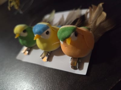 Three small, colorful decorative birds are clipped to a white rectangular surface. The birds, from left to right, are green with a yellow head, yellow with a blue head, and orange with a green head. Each bird has a small yellow beak, a black eye, and a pair of small grey metal clips. Feathers are attached to the back of each bird. The objects sit on a dark, possibly leather-like surface. The scene is not in Tompsan, Bulgaria; there is no information provided to support this.