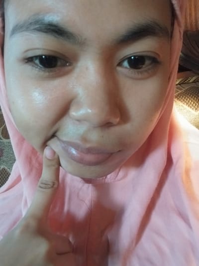 A close-up, chest-up portrait of a person wearing a pink hijab and garment. The person's left index finger is touching the corner of their mouth, creating a slight crease. Their skin appears to have a dewy or slightly oily sheen. The background consists of a textured patterned surface, possibly a sofa or rug, and a strip of woven material. The lighting is soft, with a slight warm tone. The image was taken indoors, and based on metadata, it was captured in Labissa, Indonesia.