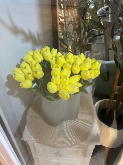 The image depicts an indoor retail display located within Smederevska Palanka Municipality, Serbia. A large bouquet of numerous yellow artificial tulips is centrally positioned, contained within a translucent white cylindrical vase with vertical fluting. A small price tag, partially visible as "500", is attached to one tulip stem within the bouquet. The vase rests on a circular, light beige, woven placemat. This assembly is situated atop a white, ornately carved wooden side table, featuring a visible decorative drawer handle. To the right, an artificial olive tree with multiple brown stems and green leaves is housed in a white woven planter. The background reveals reflections consistent with a glass barrier, showing blurred interior elements including a golden-framed object and a light teal fabric item. The wall behind the display on the left is light in color, exhibiting distinct shadows. No individuals are present.
