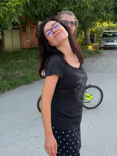 Two women are pictured outdoors on a paved road, with a bicycle wheel visible between them. The woman in the foreground, facing right and slightly away from the camera, has dark hair and is wearing a black t-shirt with a graphic print and a black skirt with white polka dots. She has on glasses with purple frames and is looking upwards and to her left. The second woman, partially obscured behind the first, has blonde hair and is wearing glasses with dark frames. She is looking forward. In the background, a silver car with a license plate visible (EM 6617 KP) is parked on the right side of the road. To the left of the car, a dark trash bin is situated. A building with a red facade and an air conditioning unit on the wall is visible behind the women, with trees and greenery surrounding the area. The overall setting appears to be a residential street in the city of Dolni Dabnik, Bulgaria.