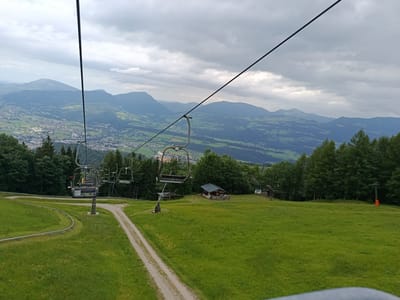 The image depicts a view from a ski lift in Hallein, Austria. The foreground is dominated by a lush green grassy slope dotted with yellow wildflowers. A gravel path winds its way up the slope. In the middle ground, a ski lift with several empty chairs is visible, supported by a sturdy pole. To the right of the ski lift, a small, rustic wooden cabin with a dark roof sits in the grass. Beyond the cabin, a dense line of dark green coniferous trees forms a wall. The background reveals a vast panorama of rolling hills and mountains stretching into the distance under a cloudy sky. A town with scattered buildings can be seen nestled in the valley below the mountains, suggesting the location is on the outskirts of a populated area. The overall scene is serene and natural, with a hint of human activity from the ski lift and the cabin.