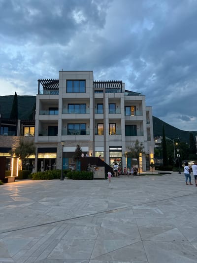 The image shows a modern, multi-story building with a pale facade and numerous balconies, situated on a paved plaza. Several people are visible on the plaza, some walking and others gathered near the building, which appears to house shops or restaurants. The building's balconies have glass railings and some have wooden louvre structures above them. The lower levels of the building feature illuminated shopfronts.
In the background, a lush green mountain slopes upwards, its peak partially obscured by dramatic, overcast clouds. The sky is a mix of dark grey and lighter patches, suggesting dusk or a stormy evening. The paved plaza is laid with large, light-colored stone tiles arranged in a geometric pattern.
A few trees, including tall cypresses, are planted along the edge of the plaza and in front of the building. Streetlights are also visible, casting a warm glow on the plaza and the building's facade.
The overall scene conveys a sense of a bustling public space during twilight, with a backdrop of natural beauty. The time of day is clearly evening, evidenced by the artificial lighting and the moody sky. The location context is Kumbor, Montenegro, as indicated.
Visible text includes "MEMADE" and "FRESHING" on illuminated signs.