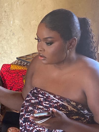 A Black woman with dark skin and makeup is shown from the chest up, looking to her left with her mouth slightly open. She has her hair pulled back in a high ponytail. She is wearing a colorful patterned fabric wrapped around her. To her left, red and yellow patterned fabric is visible, and behind her, a textured, light-colored wall can be seen. A dark tray with circular indentations is also partially visible. The scene appears to be indoors, possibly in a domestic setting in Panyam, Nigeria.