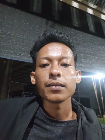 This is a selfie taken by a young man outdoors at night. He is wearing a black jacket over a dark grey t-shirt. The background is dark with some out-of-focus structures visible, including what appears to be some sort of wooden roofing or shelter. There are bright lights in the background, suggesting artificial illumination. The man's face is well-lit, highlighting his skin which appears slightly oily or sweaty. He has short, dark, styled hair and a faint mustache and goatee. His expression is neutral. The time of day is clearly night due to the lighting and darkness. The location context is Keudematangkuli, Indonesia, but there are no specific visual cues in the image to confirm this. There is no visible text in the image.