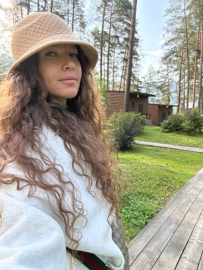 A woman wearing a woven hat and a white sweatshirt stands in an outdoor setting with trees and wooden cabins. She has long, curly brown hair and is looking towards the camera. The background features several wooden structures, lush green grass, and a wooden walkway. The scene appears to be set in Barangol, Russia, given the pine trees and rustic cabins often found in that region. The lighting suggests it is daytime, with the sun casting shadows.