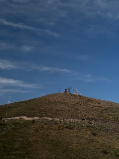 This media file captures a sunlit outdoor scene, possibly taken on a clear day with a few wispy clouds. The foreground is dominated by a grassy hillside, sloping upwards towards the right of the frame. The grass appears dry and brownish, suggesting a warm season. A faint path or track can be seen winding its way up the hill.
In the mid-ground, a series of tall, dark poles with wires strung between them are visible, indicating the presence of a power line or some other form of overhead infrastructure. Further up the slope, there are some complex structures that resemble industrial equipment, possibly related to mining or some form of extraction, with metallic frameworks and what might be cranes. These structures are silhouetted against the bright blue sky.
The sky itself takes up a significant portion of the left side of the image and is a vibrant blue, streaked with thin, white clouds. The overall impression is one of a vast, open landscape under a bright sky. There is no visible text in the image. The scene appears to be remote and undeveloped, with the natural landscape being the primary feature, interspersed with man-made structures.