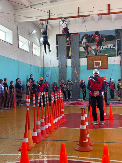 This image captures an indoor rope climbing or technical mountaineering competition within a gymnasium in Tayshet, Russia. Two individuals are suspended from an overhead orange metal beam structure near the white ceiling, maneuvering on ropes while wearing helmets and climbing harnesses. One is dressed in dark athletic wear with blue and white accents, the other in a white top and dark pants.
A tall, grey climbing wall is visible on the far wall, positioned beneath a large mural depicting two soccer players and a "WATTYL" sign. A wooden basketball backboard with a hoop and net is also mounted on this wall. The polished wooden floor features red, yellow, and blue court markings. A long line of orange and white striped plastic cones, interspersed with smaller solid orange cones, delineates a path or activity zone across the foreground and midground.
Several individuals wear red vests with "ФСТР СУДЬЯ" (FSTR Judge) printed in white Cyrillic script. One judge stands in the foreground with their back to the camera, hands clasped behind them. Other judges, some wearing orange helmets, are dispersed around the event area. On the left, a group of approximately 15 observers or participants, some in climbing gear, stand behind a dark safety net, looking upwards at the suspended climbers. Ropes are coiled on the floor in various locations. The gymnasium features light blue lower walls, white upper walls, and multiple rectangular windows on the left side, along with overhead fluorescent lighting fixtures. A blue flag with a white emblem hangs from the upper left wall.