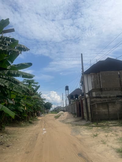 The image depicts a dirt road in Rumuosi, Nigeria, lined with lush green banana trees on the left and ongoing construction on the right. The sky is partly cloudy with scattered white clouds against a bright blue backdrop. On the right side of the road, a multi-story building under construction is visible, with its roof covered in dark shingles and walls made of concrete blocks. A tall water tower stands in the distance behind a fence. The road itself is unpaved and shows tire tracks, with some puddles of water scattered on its surface. A pile of sand is also present near the construction site. The overall scene suggests a developing area with residential and construction activity.