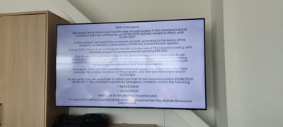 A television screen is displaying a message to colleagues regarding a new insurance plan commencing on 01/06/2026 and remaining in effect until 31/05/2027. The notice explains a mandatory deductible of €100, increased insurance usage leading to premium adjustments, but that the company will absorb the cost for both primary insured members and their dependents. It notes that the affiliated hospitals for emergency incidents, Agios Loukas and Geniki Kliniki, remain the same. The message concludes with a request to contact the Human Resources Department for clarification. The screen is positioned next to a light brown cabinet, and what looks like a black bag is underneath. The setting is Λάκκωμα, Greece.