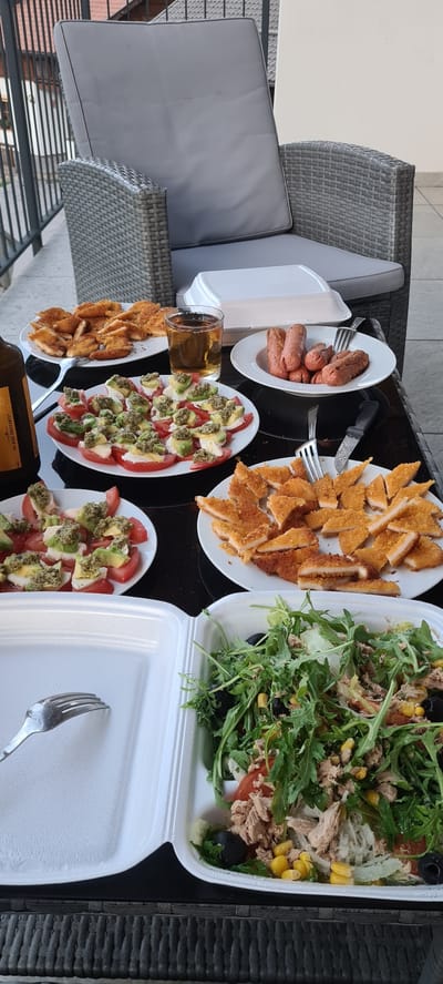 A table laden with food sits outdoors on a patio, likely in Deutschnofen - Nova Ponente, Italy. The table, with a black glass surface and a woven rattan base, displays a spread of dishes. In the foreground, a white styrofoam container holds a salad mixture of green leafy vegetables, corn, olives, and flaked tuna, with some shredded white material, possibly cheese or onion. Next to it, a metal fork rests in the empty portion of the container.

Moving further back on the table, a plate of breaded chicken pieces is arranged. Behind this, a plate of tomato and mozzarella slices topped with green pesto and avocado can be seen. To the right of the chicken, a white plate holds several cooked sausages. A glass of amber-colored liquid, likely beer, is positioned between the chicken and sausage plates. Further back, a bottle with a yellow label, partially obscured, is present. In the background, a grey woven patio chair with cushions is visible, suggesting a relaxed outdoor dining setting. The overall scene depicts a meal prepared for enjoyment in an outdoor environment.