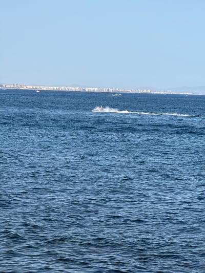 The image captures a bright, clear day at sea near the city of Aguadulce, Spain. The foreground is dominated by the deep blue, rippled surface of the Mediterranean Sea, with small waves creating a textured pattern. In the mid-ground, a person is riding a jet ski, leaving a white wake behind them as they move across the water. Further in the distance, a long, low-lying coastline is visible, lined with numerous buildings that make up the city of Aguadulce. A few distant boats and smaller vessels can be seen dotting the sea near the shore. The horizon is marked by a clear, pale blue sky, with a faint haze obscuring the distant mountains. The overall scene is one of recreational activity by the coast under good weather conditions.