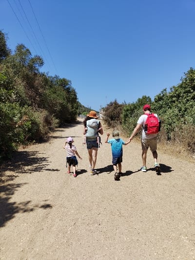 The image captures four individuals walking away from the camera on a wide, unpaved dirt path under a clear blue sky in the Maale Yosef Regional Council, Israel. The path, light brown with visible gravel, is flanked on both sides by dense green and brown shrubbery and trees. From left to right, the subjects are a young girl, an adult female, a young boy, and an adult male.
The young girl, positioned on the far left, wears a light pink cap, a light-colored t-shirt, and a dark patterned skirt. The adult female, located to the right of the girl, wears a light-colored top, dark shorts, and a light brown wide-brimmed hat. She carries an infant or small child in a front-facing carrier on her chest and also has a light-colored backpack. The young boy, to the right of the adult female, wears a blue ombre t-shirt and dark shorts. He holds the right hand of the adult male. The adult male, on the far right, wears a white t-shirt, light shorts, a red baseball cap, and a red backpack.
Shadows are cast from the left onto the foreground of the dirt path. In the upper left background, multiple power lines are visible traversing the clear blue sky. A faint, light-colored structure is partially discernible through the distant vegetation on the horizon. The scene is illuminated by direct sunlight.