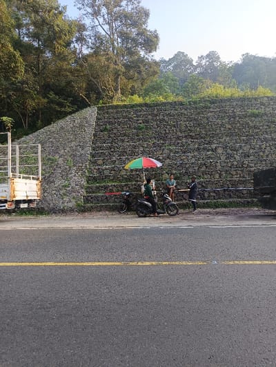 This image depicts a roadside scene in Cugenang, Indonesia, under clear daylight conditions.
In the foreground, an asphalt road with a solid yellow center line and a broken white shoulder line stretches horizontally across the lower third of the frame. The road appears dry and well-maintained.
On the dirt and gravel shoulder to the right of the road, a stepped gabion retaining wall, constructed from wire mesh filled with rocks, dominates the midground and background, ascending towards a densely forested slope. The wall is multi-tiered, with small patches of green vegetation growing within its structure.
Three individuals and several vehicles are present on the shoulder, positioned in front of the retaining wall. From left to right along the shoulder:
* A white flatbed truck with a metal cage structure is partially visible on the far left, parked near the base of the retaining wall.
* Two black scooter-style motorcycles are parked adjacent to each other. One person, dressed in a dark long-sleeved shirt and dark pants, is seated on the rightmost scooter. A small, open umbrella with red, yellow, and green segments is positioned directly in front of this person, suggesting a roadside vendor setup.
* A second person, wearing a light blue t-shirt and dark shorts, is seated on a low structure (possibly a stool or crate) to the right of the vendor, facing them.
* A third person, dressed in a black long-sleeved top with white arm stripes and dark pants, stands to the right of the seated individual, engaging in apparent conversation with them or the vendor.
* A dark-colored truck is partially visible on the far right, parked on the shoulder.
The background consists of a dense forest of tall trees and lush green foliage covering the upper portion of the slope above the retaining wall, under a clear, light blue sky.