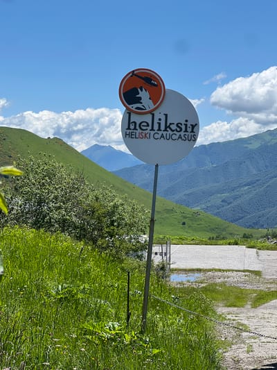 The image depicts a sign for "Heliski Caucasus" set against a backdrop of lush green mountains under a bright blue sky dotted with white, fluffy clouds. The sign itself is circular with two parts. The top part is a smaller orange circle with a black silhouette of a helicopter flying over a mountain peak. The bottom part is a larger white circle with the text "heliksir" in black, stylized font, and below that, "HELISKI CAUCASUS" in smaller black capital letters.
The sign is mounted on a metal pole in a grassy area with tall green grass and some wildflowers. To the right of the sign, a paved area is visible, part of which is covered by shallow puddles of water. In the background, rolling green hills and more distant, hazy blue mountains create a sense of depth. A few trees are scattered on the hillsides. The overall impression is one of a remote, mountainous natural landscape, likely a location for outdoor adventure activities. There are no people visible in the image. The location is identified as Kumlistsikhe, Georgia.