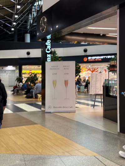 The image captures a view of the Mercedes-Benz Cafe at Sheremetyevo International Airport (SVO) in Perepechino, Russia. In the foreground, a poster displays two glasses of sparkling wine, one white labeled "PROSECCO" and one rose labeled "ROSE," with prices listed below. To the right of the poster, a display cabinet contains bottles of alcoholic beverages. In the background, several people are seated in a waiting area, with luggage nearby. The cafe's signage is visible above an entrance. The floor is a combination of wood-patterned tiles and grey stone tiles, with diagonal black and white stripes.
