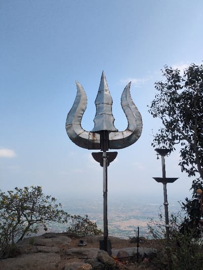 A large, silvery-metallic Trishula (trident), indicative of Hindu religious symbolism, stands prominently on a rocky elevated outcrop. The Trishula is mounted on a dark pole, with additional smaller, dark metallic pole structures featuring circular platforms and a bell visible to its right. The ground immediately surrounding the base is composed of dark grey rocks and sparse green and yellow-flowered vegetation. In the background, a vast, hazy panoramic view of a distant urban or rural landscape with fields and structures stretches under a clear, light blue sky with minimal cloud cover. Dark green trees partially frame the scene on the left and right. The scene is located in the city of Kadaladi, India. No individuals are present.