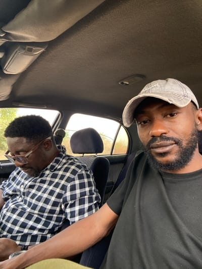 Inside a vehicle in Ungwan Liman, Nigeria, two adult males are seated in the front cabin during daylight hours. The individual in the foreground, positioned on the right side of the frame, is wearing a faded grey baseball cap and a black t-shirt, looking directly at the camera. He has a dark beard and mustache. To his left, the second individual, presumably the driver, is wearing eyeglasses with brown frames and a white and dark-colored plaid long-sleeved shirt. He has dark, curly hair and is looking downwards, appearing to interact with an object held in his hands, partially obscured below the frame. The car's interior features a light grey textured headliner, a dome light fixture, and dark grey patterned seat upholstery. Through the rear windows, sparse vegetation and a dusty or hazy outdoor environment are visible. The scene suggests a stationary or moving vehicle on a road.