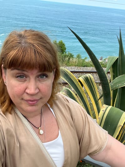 This is a portrait of a woman with auburn hair and green eyes, smiling at the camera. She is wearing a light brown jacket over a white top and a necklace with a circular pendant. The background features a body of water, possibly the sea, with a lush green plant with striped leaves, likely an agave, in the foreground. In the middle ground, there are railway tracks and some vegetation. The lighting suggests it is daytime, and the weather appears to be fair. The overall impression is one of relaxation and enjoyment of an outdoor setting, possibly a coastal location like Tsandrypsh, Abkhazia, given the presence of the sea and the type of vegetation. The image is oriented vertically, with the woman's face close to the viewer.
